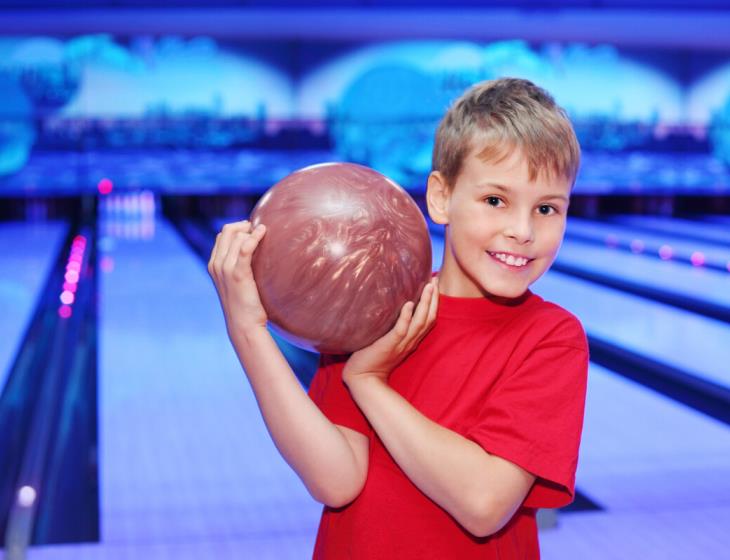 Little smiling boy dressed in red T-shirt holds ball in bowling club